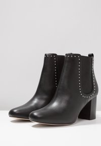 Černé kožené kotníkové boty s špičatou špičkou a blokovým podpatkem, zdobené stříbrnými nýty podél elastických bočních panelů. Hladká textura.