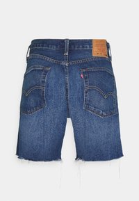 Shorts en denim bleu moyen avec un ourlet effiloché, comprenant deux poches arrière et une étiquette en cuir beige à la taille. Design classique à cinq poches.