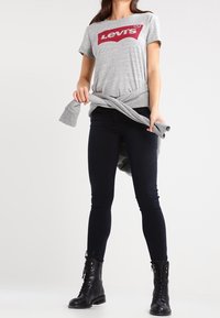 T-shirt gris à manches courtes avec logo Levi's rouge, associé à un jean skinny noir et des bottes de combat à lacets noires. Détail de nœud à la taille.
