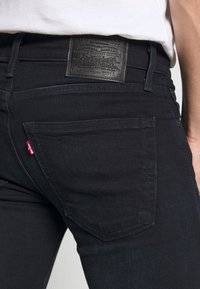 Svarta skinny jeans har en lädermidjepatch, subtila sömnadsdetaljer och en logotag på bakfickan. Slät textur, åtsittande design.