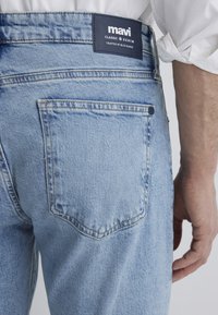 Light blue denim jeans with a back pocket and branded leather tag. The fabric features a faded texture and contrast stitching.