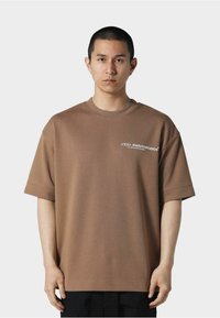 Brun oversized t-skjorte i myk bomull, med rund hals og korte ermer. Frontgrafikk i hvit tekst. Enkelt bakdesign.