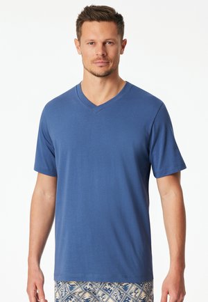 Mann, der ein schlichtes blaues V-Ausschnitt-Kurzarmshirt und gemusterte Shorts trägt, steht vor weißem Hintergrund.