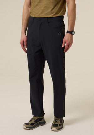 Hombre vistiendo pantalones negro para exteriores con un logo, camisa verde oliva, reloj negro y zapatos de senderismo beige, de pie sobre un fondo liso.