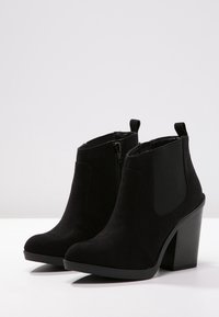 Bottines en daim noir avec un talon bloc haut, des panneaux latéraux élastiques et une fermeture éclair centrale. Surface lisse avec un design à bout arrondi.