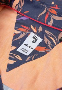 Stof met een bloemenpatroon op navy blauwe basis en oranje tinten, met een wit label waarop "J&Joy" en "Ontworpen in België" staat.