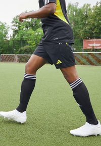 Pantalones cortos deportivos negros y calcetines negros con rayas blancas, combinados con zapatillas de fútbol blancas, en un campo de césped artificial verde.