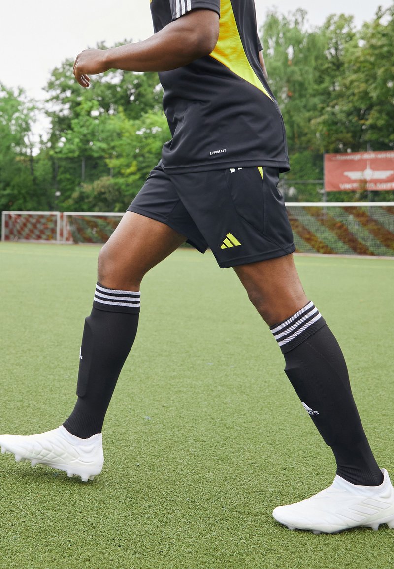 Pantalones cortos deportivos negros y calcetines negros con rayas blancas, combinados con zapatillas de fútbol blancas, en un campo de césped artificial verde.