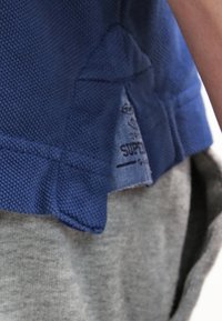 Ourlet de chemise en tissu bleu texturé avec une petite étiquette visible portant la mention «SUPER» sur un pantalon gris en tissu doux.