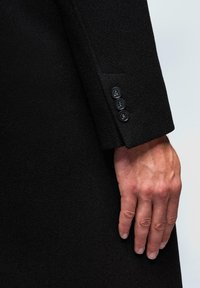 Nahaufnahme einer Hand, die neben dem Ärmel eines schwarzen Mantels mit drei schwarzen Knöpfen an der Manschette ruht.