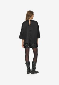 Sort satin top med brede ærmer, med en rygudskæring der er bundet med en sløjfe. Parret med sorte paillet-shorts og skinnende knæhøje støvler.