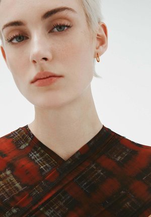 Top envuelto rojo con estampado y escote en V, que presenta tonos negros y terracota, combinado con un pendiente de aro dorado. Textura textil suave.