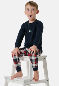 Navyblauer Langarm-Pyjama-Oberteil mit Design von Bergbäumen, kombiniert mit rot-weiß-blau karierten Pyjamahosen, sitzt auf einem weißen Hocker.