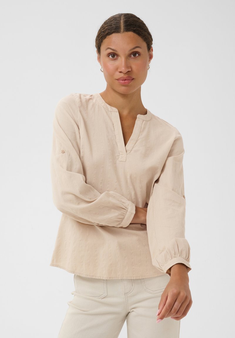 Femme aux cheveux tressés portant un chemisier beige ample à manches longues et un pantalon crème, debout avec un bras croisé et l'autre détendu.