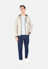 Giacca impermeabile color beige chiaro con cappuccio, cerniere nere e polsini. Indossata su una maglia grigia e abbinata a jeans blu e sneaker bianche.