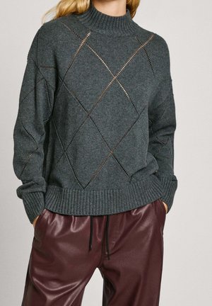 Femme portant un pull en maille gris foncé avec motif ajouré en losange et un pantalon en cuir bordeaux avec cordon noir.