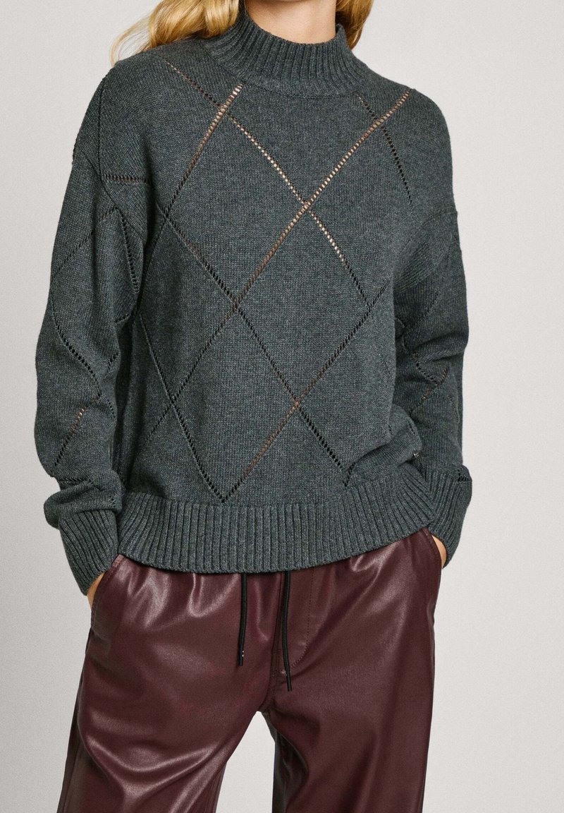 Femme portant un pull en maille gris foncé avec motif ajouré en losange et un pantalon en cuir bordeaux avec cordon noir.