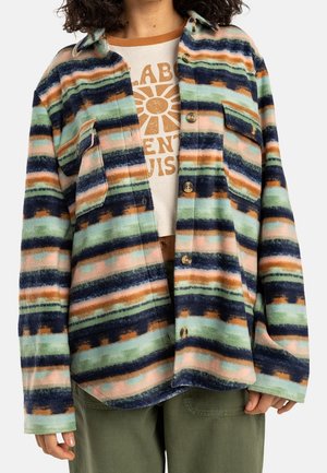 Person iført en knappet, stribet fleecejakke i grøn, marineblå, orange og pink over en beige grafisk T-shirt med brun tekst og olivengrønne bukser.