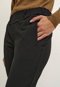 Pantalon noir avec une texture lisse, taille élastique, poches latérales et une coupe ajustée. Le tissu semble extensible et près du corps.