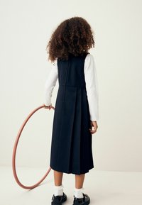 Navy-blaues Faltenkleid mit Reißverschluss im Rücken, kombiniert mit einem weißen Langarmshirt. Schwarze Schuhe und weiße Knöchelsocken runden den Look ab.