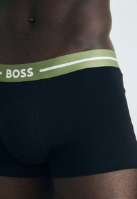 Boxer neri con una fascia elastica verde che presenta la parola "BOSS" in bianco e una sottile striscia bianca, realizzati in un tessuto morbido.
