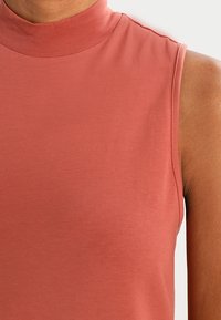 Ärmlös topp i mjuk terrakottafärg med hög halsringning och slät textur, med en figurnära design som följer kroppens former.