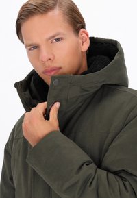 Jachetă verde măslinie cu glugă căptușită cu fleece, închidere cu capse și țesătură texturată. Are un guler înalt și detalii cusute.