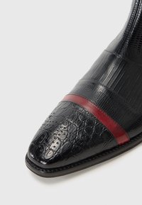 Sorte dressko med teksturert lær og spiss tå, akseptert med en burgunderstripe. Glatt, blank overflate med detaljerte mønstre.