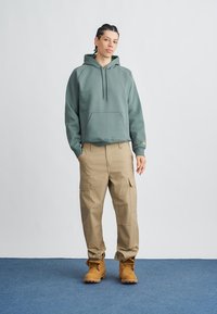 Zelena majica s kapuljačom s prednjim džepom, opušteni kroj; kaki cargo hlače s više džepova; smeđe čizme. Jednostavan dizajn, mekana tekstura.