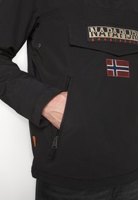 Chaqueta de pullover negra con un bolsillo frontal, presenta un parche con la bandera noruega y un logotipo bordado. Bolsillo lateral con cremallera visible.