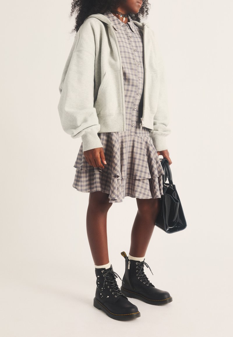 Personne portant une robe à carreaux, un sweat à capuche zippé clair, des bottes noires à lacets, des chaussettes blanches et tenant un sac à main noir.