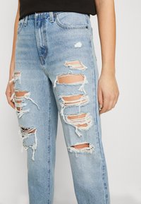 Ljusblå denimjeans med stora slitna hål på framsidan av låren och knäna, bärs med en svart topp, hand vilar vid sidan.