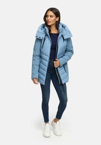 Light blue puffer jacket with a detachable hood, zipper front, and button accents; paired with dark jeans and white sneakers.
