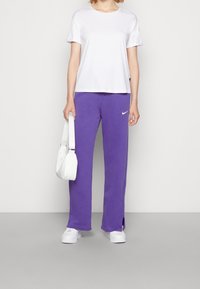 Biała koszulka z krótkim rękawem i okrągłym dekoltem, w zestawie z fioletowymi spodniami dresowymi z dwoma bocznymi rozcięciami; trzyma białą torebkę; białe sneakersy.