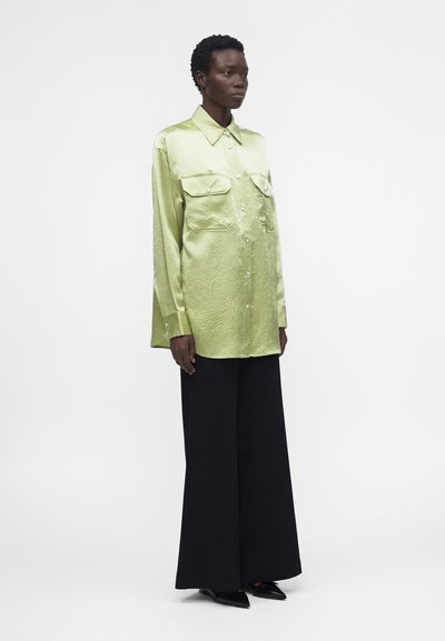 Γυναίκα που στέκεται φορώντας lime πράσινο σατέν πουκάμισο με μακριά μανίκια και τσέπες με καπάκι και μαύρο παντελόνι με φαρδιές μπατζάκια πάνω σε απλό λευκό φόντο.