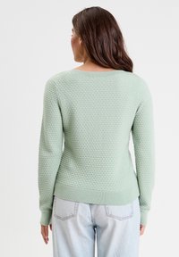 Lys grøn tekstureret sweater med et honningkage mønster, rund halsudskæring og ribbede manchetter og kant, båret med lyseblå jeans.