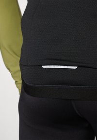 Gros plan sur une personne portant un pantalon de sport noir et un haut noir avec une petite étiquette logo réfléchissante près de la taille.