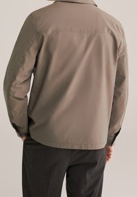 Uomo che indossa una camicia a maniche lunghe color marrone chiaro con polsini abbottonati e pantaloni grigio scuro, con le mani parzialmente nelle tasche, visto da dietro.