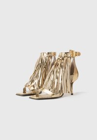 Gold high-heeled sandals with fringed detailing, open-toe design, and ankle strap. Made of metallic material with a shiny finish.