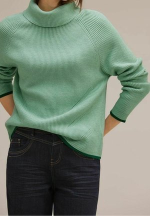Femme portant un pull à col roulé côtelé vert clair avec bordure vert foncé et un jean bleu foncé, posant les mains dans le dos.