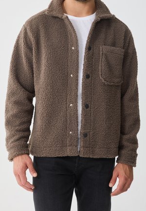 Mężczyzna w brązowej, teksturowanej kurtce polarowej z przednimi zatrzaskami i kieszenią na piersi, na białej koszuli i czarnych spodniach.