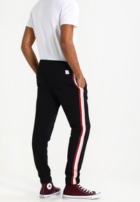 Zwarte taper joggers met rode en witte zijstrepen, elastische tailleband en vergrendelde enkels, gecombineerd met bordeauxrode hoge sneakers.