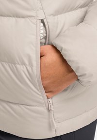 Hellgraue Steppjacke mit horizontalen Nähten, ausgestattet mit einer Reißverschlusstasche und glatter Oberfläche. Elastischer Reißverschluss-Zug an der Tasche.