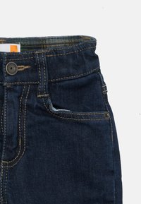 Temno modri denim jeans z zapiranjem na gumb, kontrastnim šivanjem in majhnim žepom na sprednji strani. Gladka tekstura, klasičen kroj.