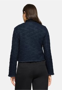 Femme aux longs cheveux bruns portant une veste texturée bleu foncé et un pantalon noir, vue de dos sur un fond uni.