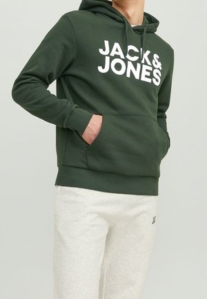 Mężczyzna w ciemnozielonej bluzie z kapturem z napisem „JACK & JONES” oraz jasnoszarych spodniach dresowych, ręce w kieszeniach bluzy.