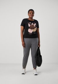 Black t-shirt with a graphic design in pink and brown, paired with gray sweatpants. Model holds a black pleated tote bag. White sneakers worn.