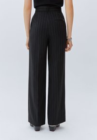 Pantalones de pierna ancha negros con rayas verticales, tejido texturizado, cintura alta, lazos para cinturón y un bolsillo trasero. Detalle de zapato liso y pulido visible.