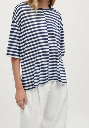 Woman wearing a loose, short-sleeve blue and white horizontally striped shirt paired with loose white pants against a plain background.