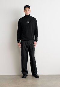 Black turtleneck sweater with logo, paired with black trousers. The model wears a matching black cap and black shoes. Simple, sleek design.